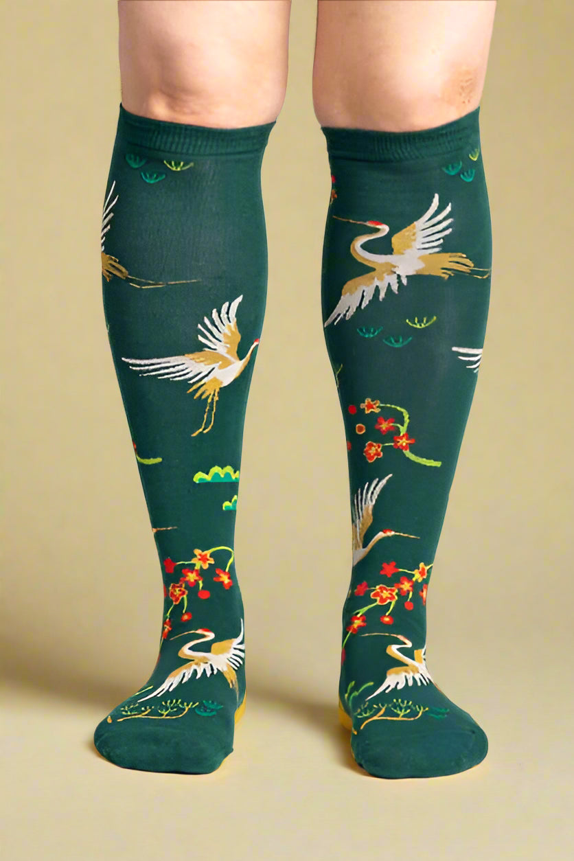 Green knee-high socks with bird and floral pattern worn by a person on a light gray background