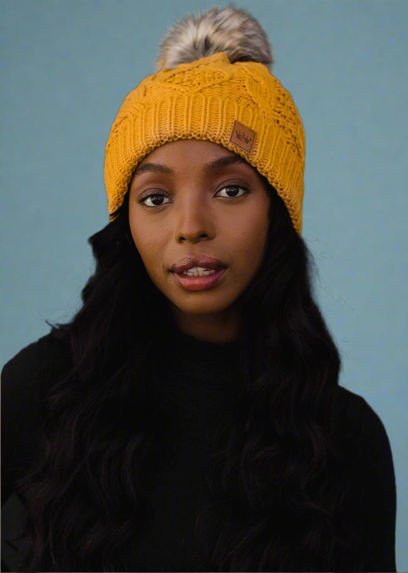 Woman wearing a yellow knit beanie with a fur pom-pom against a black background