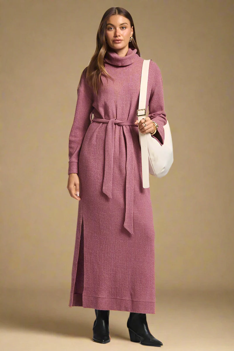 Woman wearing a long pink dress with a belt, holding a white bag on a plain background.