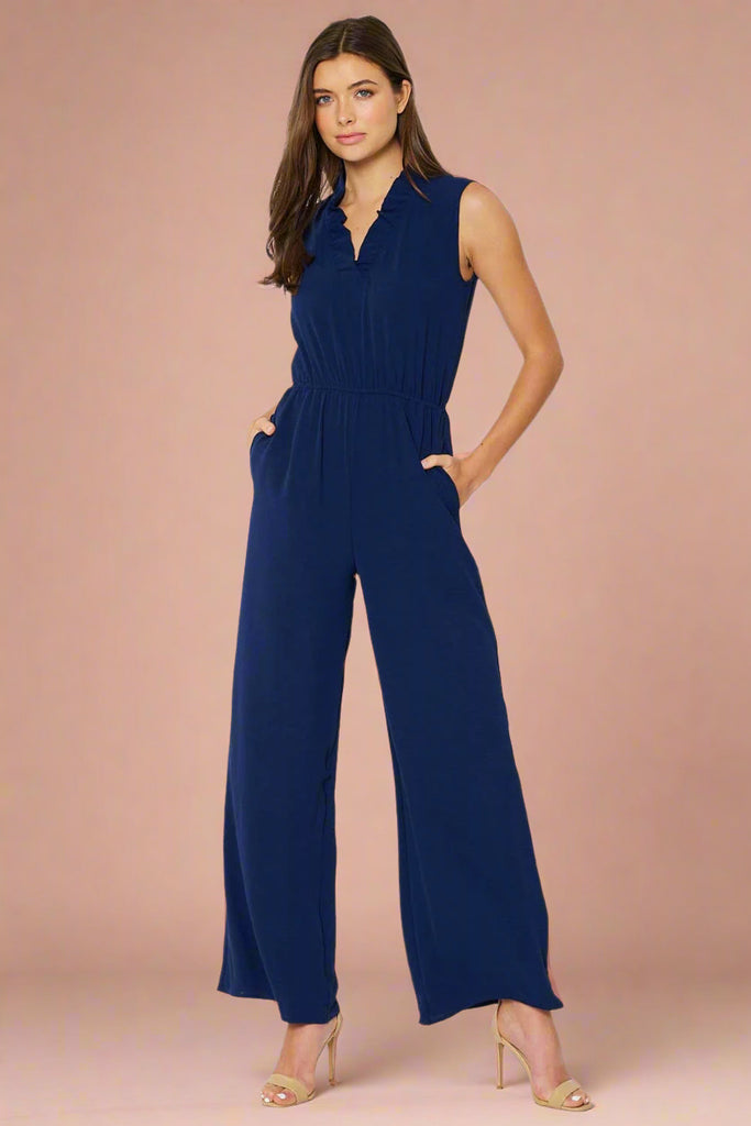 Model is wearing a flattering navy jump suit with a ruffled v neck, elastic waistband and pockets. 