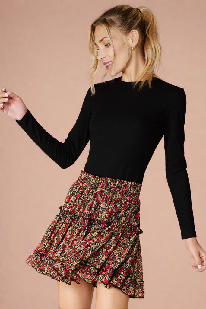 Model is wearing a floral mini skirt featuring an elastic waistband. The skirt is black with an all over floral print in red. It is fully lined and has ruffled details. 