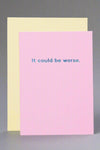 A solid pink greeting card with metallic blue text that reads ‘It could be worse.’