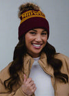 Woman wearing a maroon beanie with 'Mid West' text, tan jacket, and white shirt against a black background