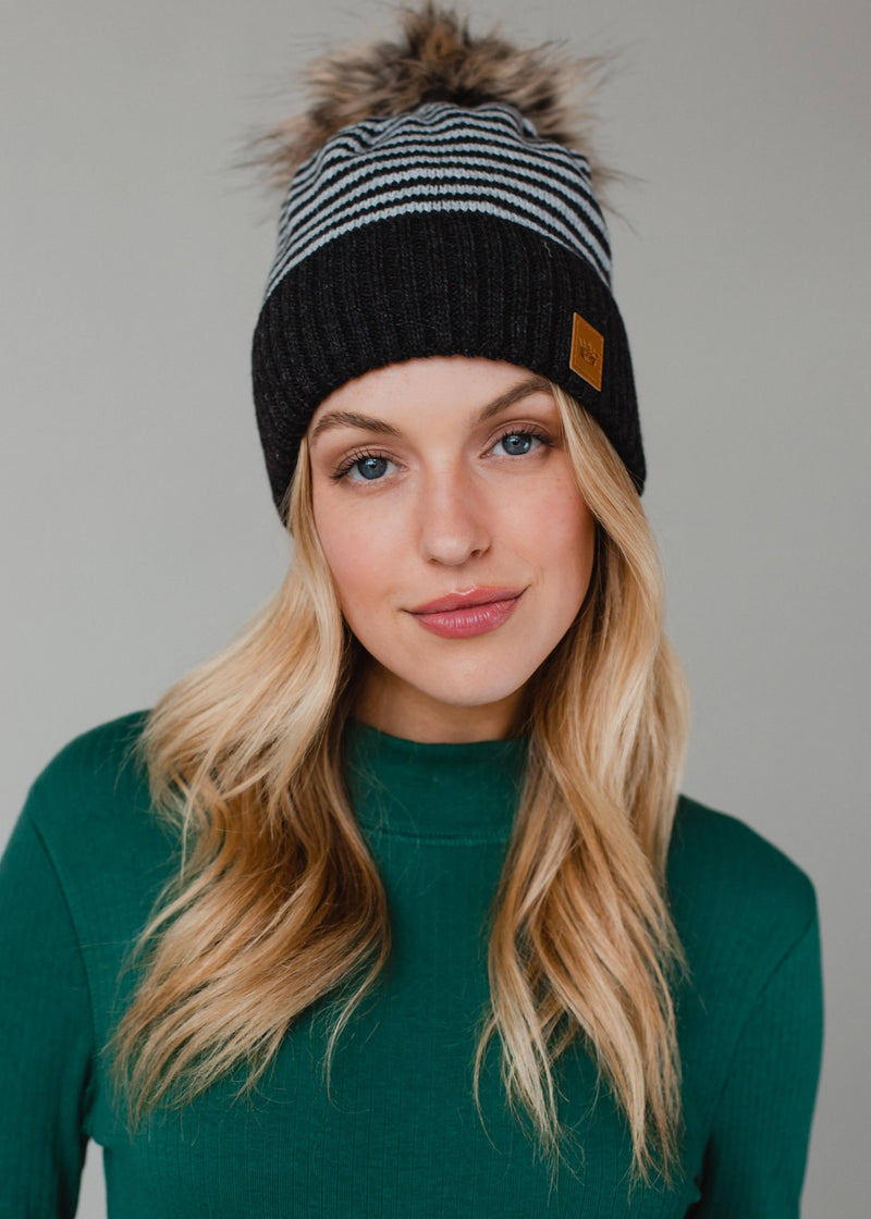 Woman wearing a black and gray striped beanie with a fur pom-pom against a neutral background