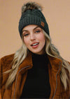 Woman wearing a dark green knit beanie with a fur pom-pom and a brown jacket on a grey background
