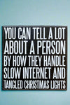 A square black sign that says “You can tell a lot about a person by how they handle slow internet and tangled Christmas lights” written in white.