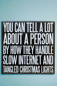 A square black sign that says “You can tell a lot about a person by how they handle slow internet and tangled Christmas lights” written in white.