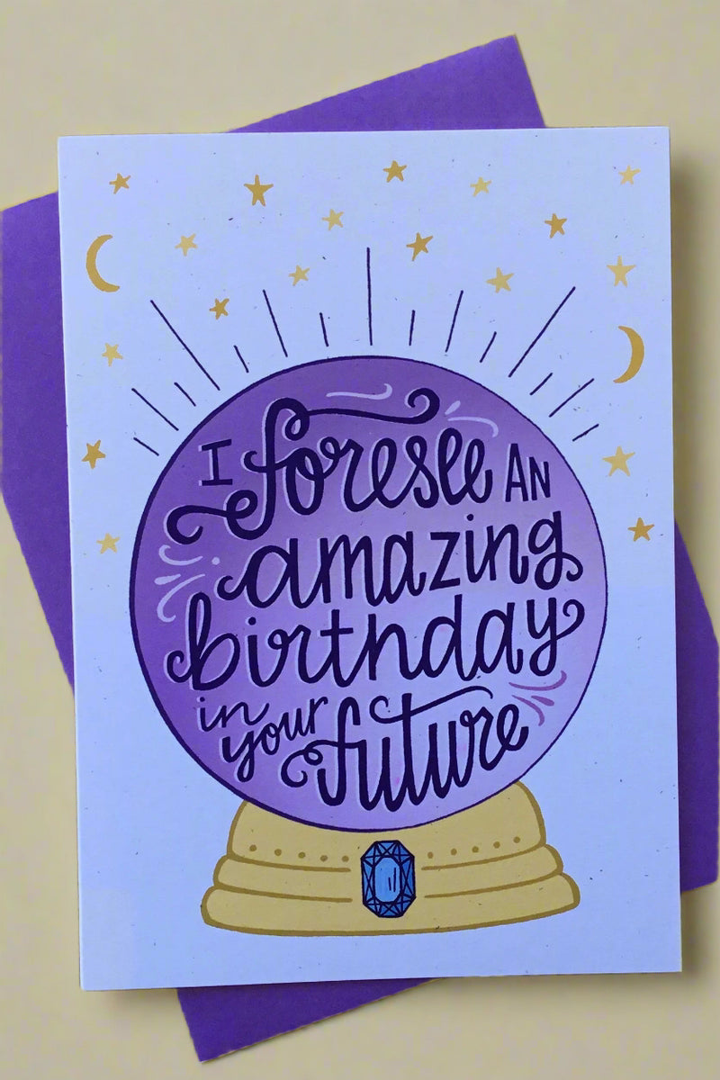 A greeting card with the image of a crystal ball on it with text in the ball that reads ‘I foresee an amazing birthday in your future’ and matching purple envelope.