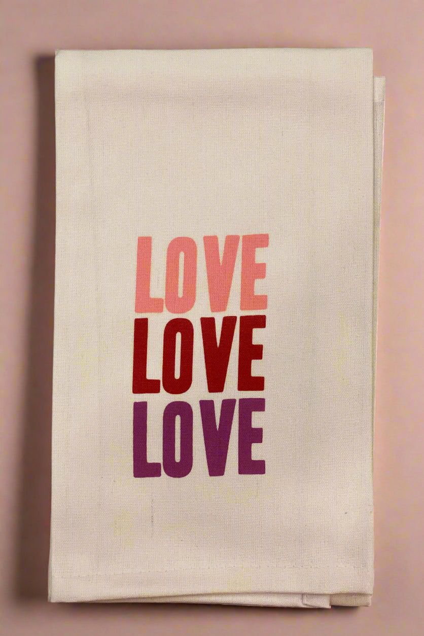 A tan, 100% cotton dish towel that has LOVE one the front in 3 different pink or red colors.