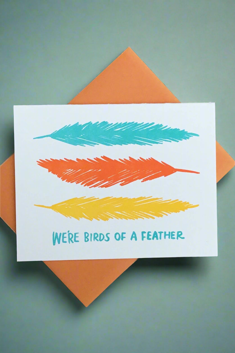 A white cord with three colorful feathers that says “we’re birds of a feather.”