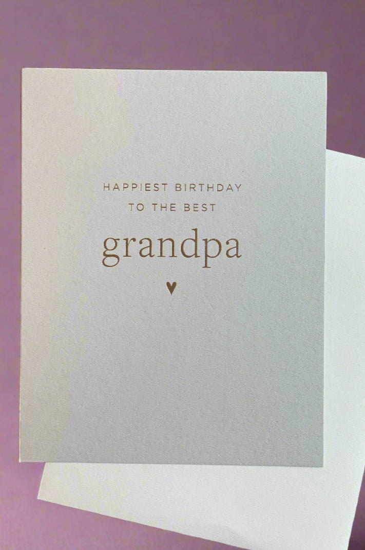 A simple white greeting card with gold text that reads ‘Happiest Birthday To The Best Grandpa’ and a white envelope.