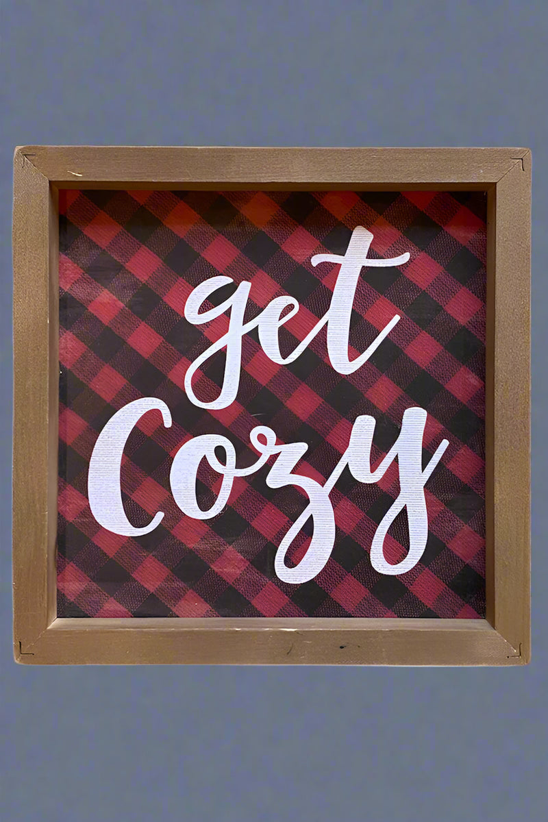 A small wood framed sign that has a red and black checkered background and “get cozy” written in white.