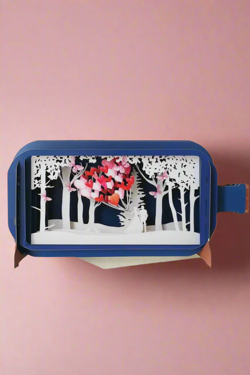 A 3D pop up greeting card in the shape of a corked bottle with a scene of a couple in the woods with heart balloons. 