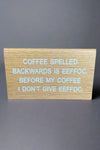 A small plastic tent sign with faux wood look and that reads ‘Coffee spelled backwards is eeffoc. Before my coffee I don’t give eeffoc.’